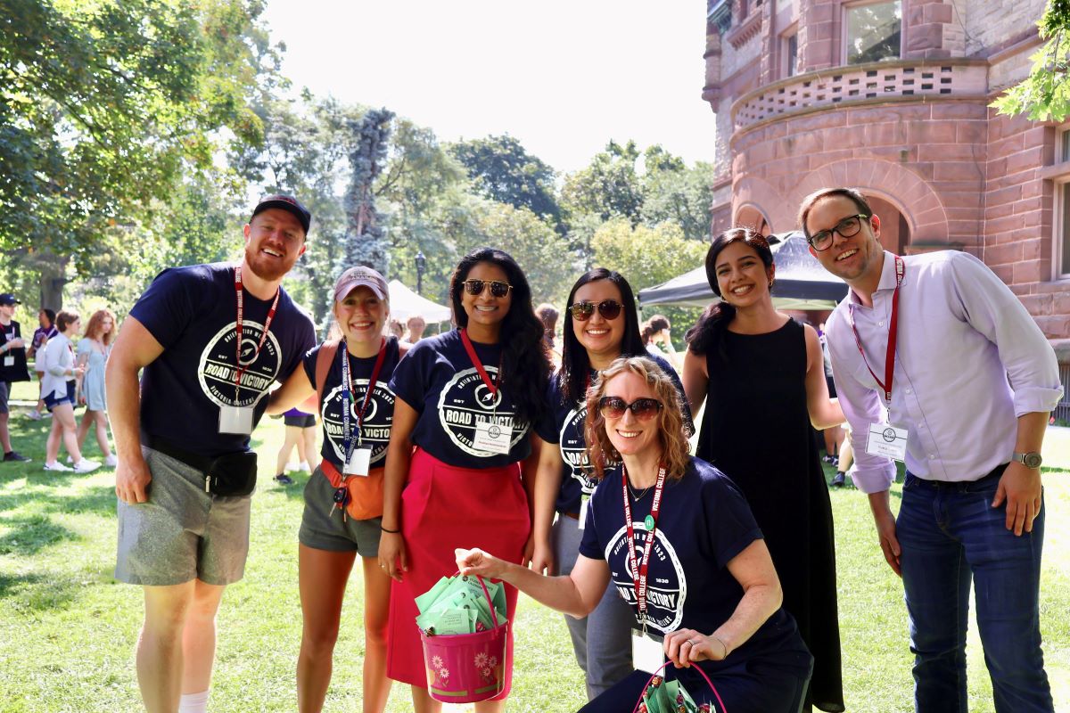 Staff members from the Dean's office gather and smile for a picture, posing in front of a lush green lawn with students strolling and mingling in the background.