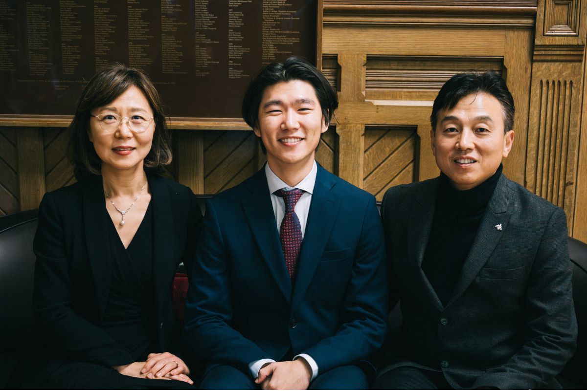 Daniel Lee sits smiling on a sofa in Victoria College, flanked by his parents.