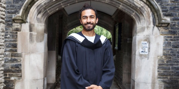 Eli Meadow Ramraj standing in front of a stone archway on the Victoria College campus in a graduation gown.