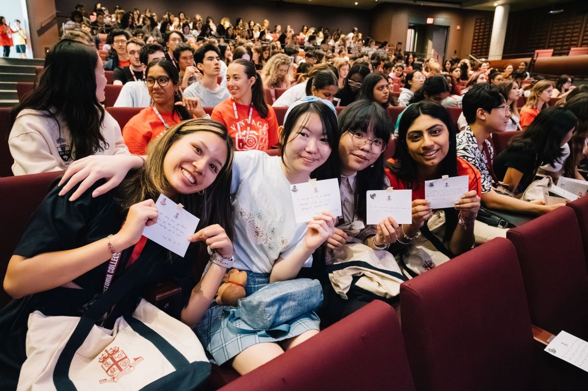 Four smiling first-year Victoria College students hold welcome messages from Vic alumni, with dozens of other students chatting in the auditorium background.