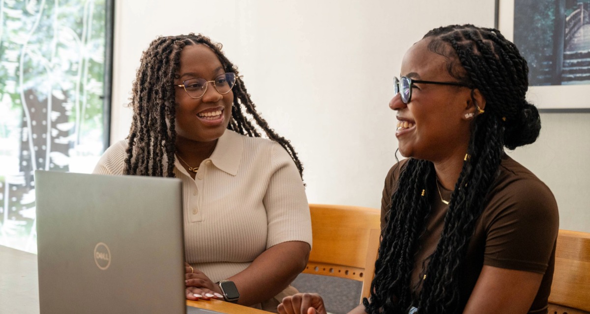 Imani King and Adura Oyedele chat while seated at a desk in front of an open laptop.