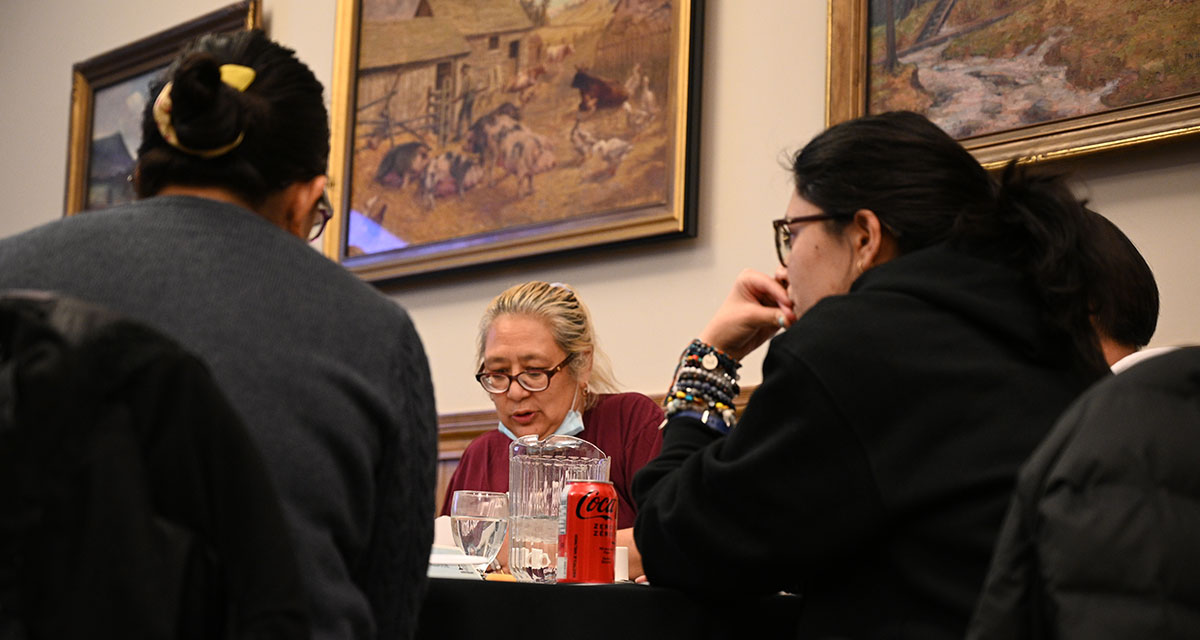 Three individuals seated at a table engage in lively conversation during a Humanities for Humanity event at Alumni Hall, Victoria University, University of Toronto.