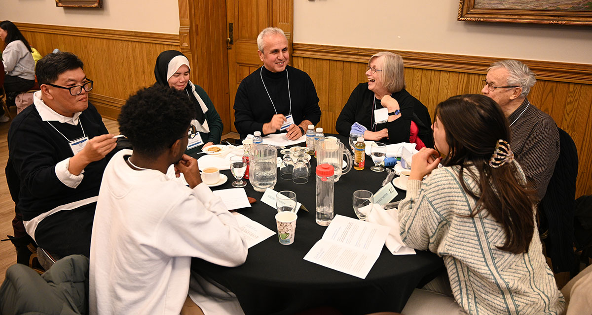 A group of seven individuals engaged in conversation and dining at a table during a Humanities for Humanity event at Victoria University, University of Toronto.